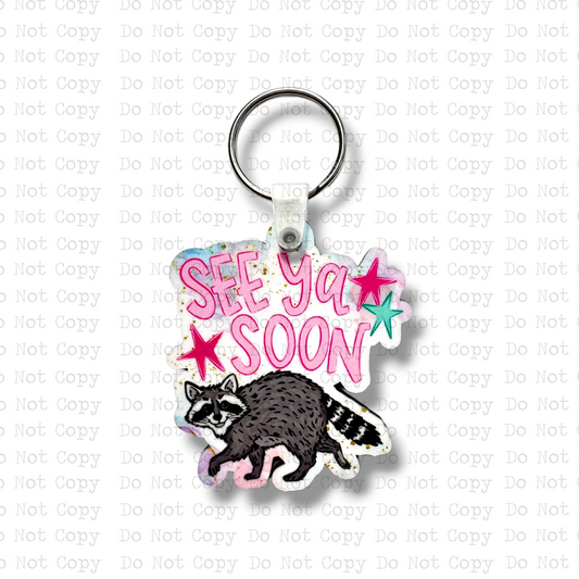 Custom-shaped contoured MDF wood sublimation keychain blank kit featuring a cute cartoon raccoon and text that says See Ya Soon with pink and teal stars, shown with keyring hardware.
